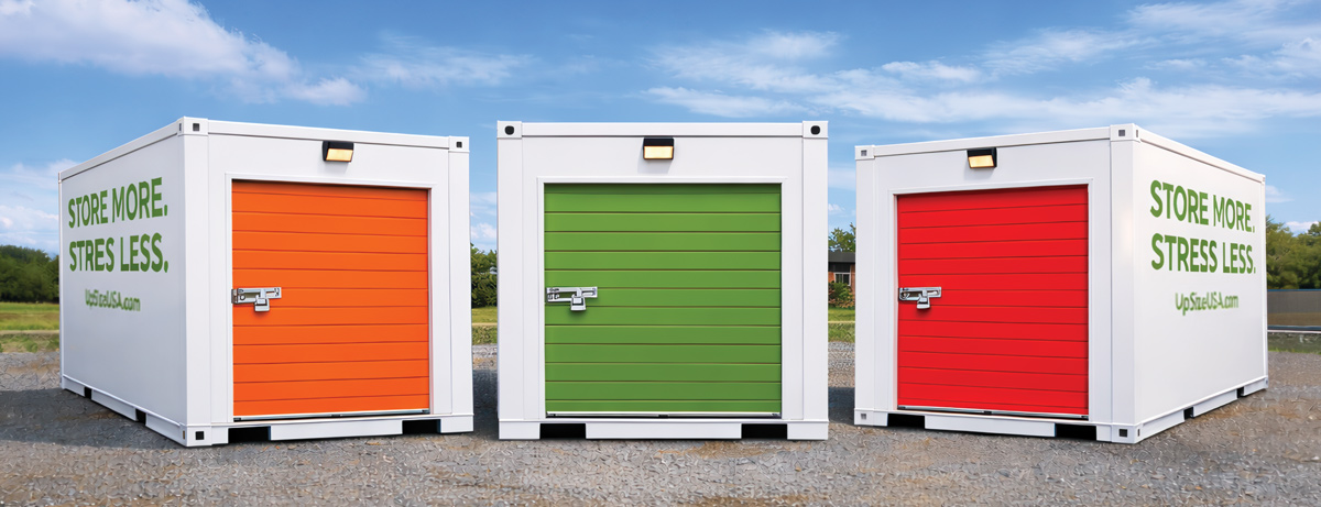 Three white "UpSize" portable storage containers with orange, green, and red roll-up doors stand in a row outdoors. The side of each unit reads, "Store More. Stress Less."