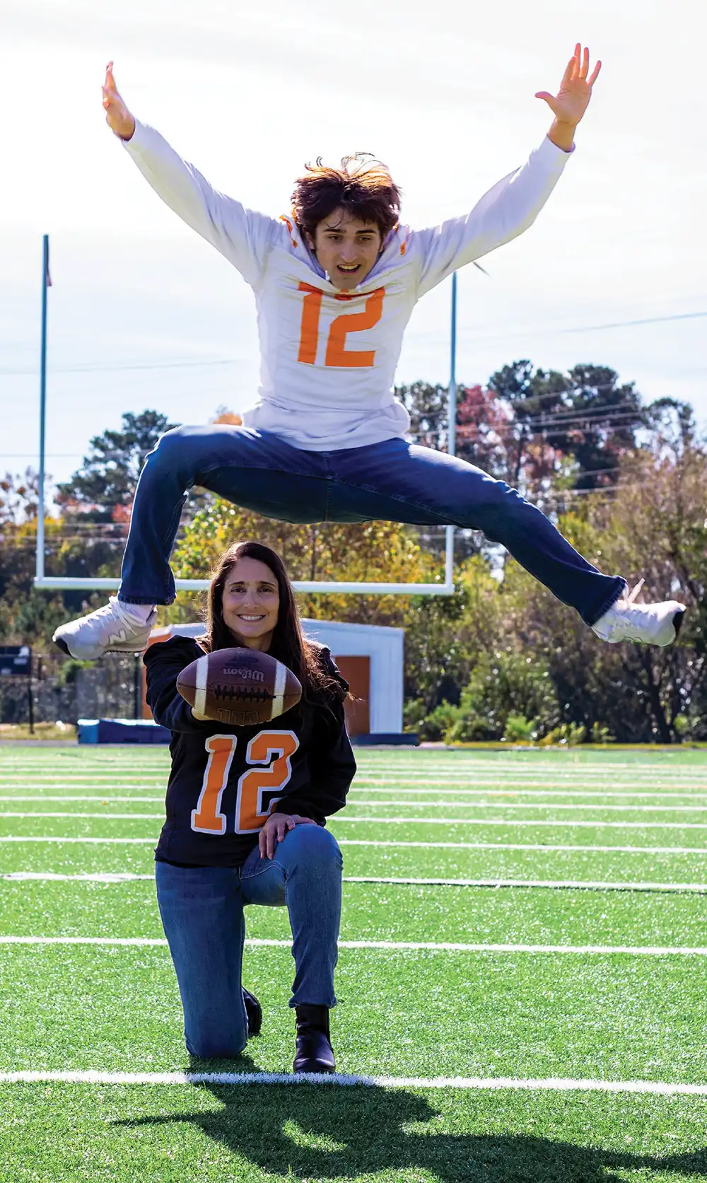 A woman in a black football jersey kneels on an athletic field holding a football, while a young man in a white jersey with the number 12 jumps high in the air behind her.