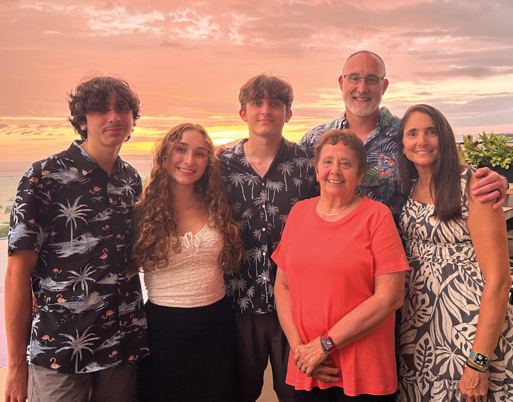A group of six family members of various ages stand together and smile for a photo outdoors during a vibrant orange and pink sunset over the water in the background.