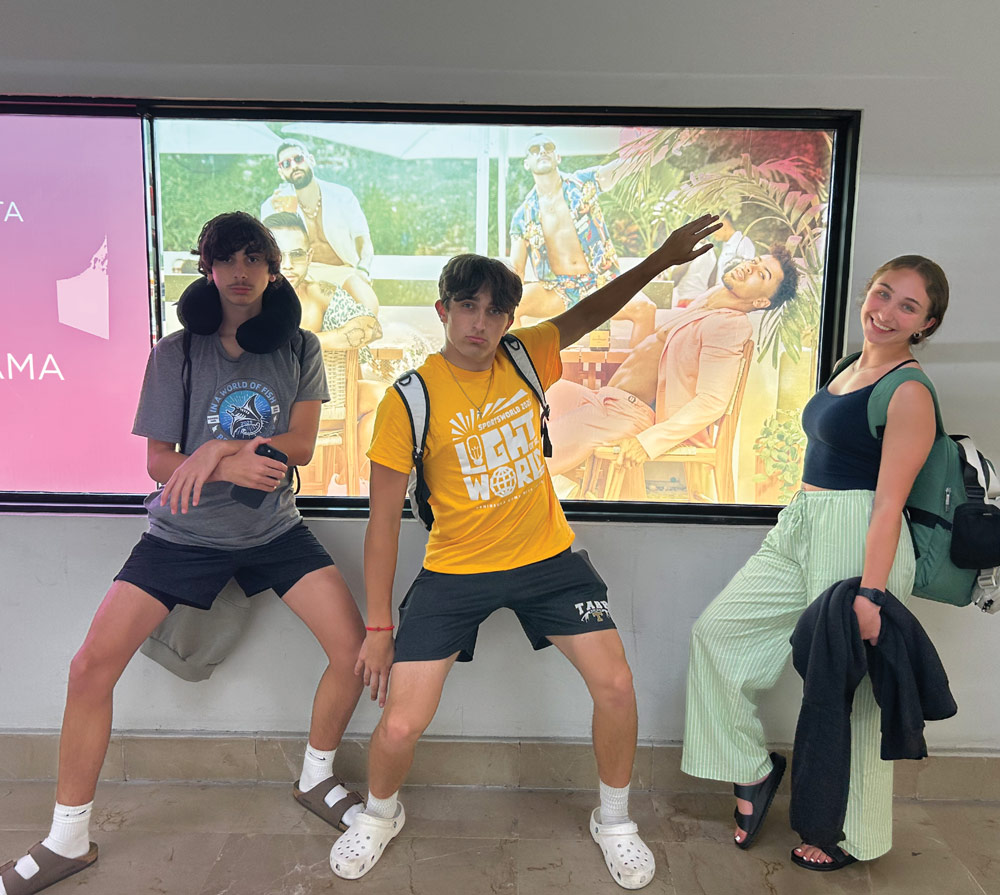Three teenagers strike playful, exaggerated poses in front of a colorful backlit advertisement in a hallway. They are dressed in casual summer clothes and wearing backpacks.
