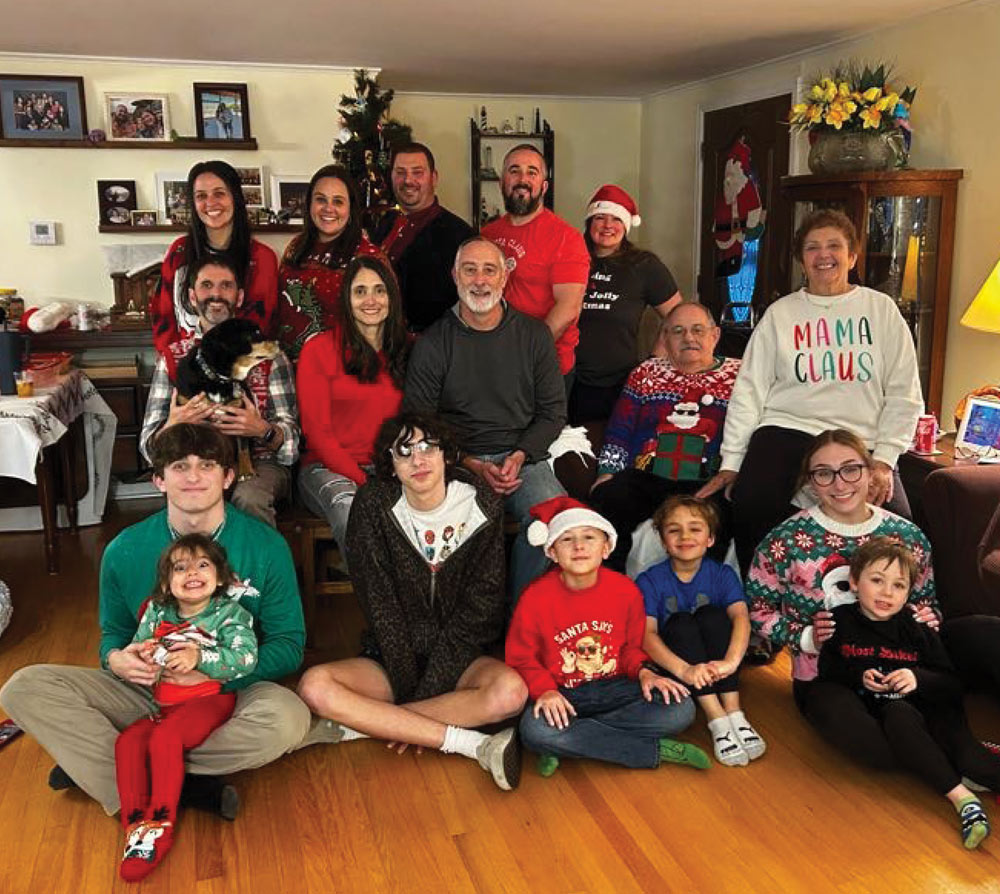 A large, multi-generational family of seventeen people and one dog pose for a group photo in a living room, with many members wearing festive Christmas sweaters and hats.