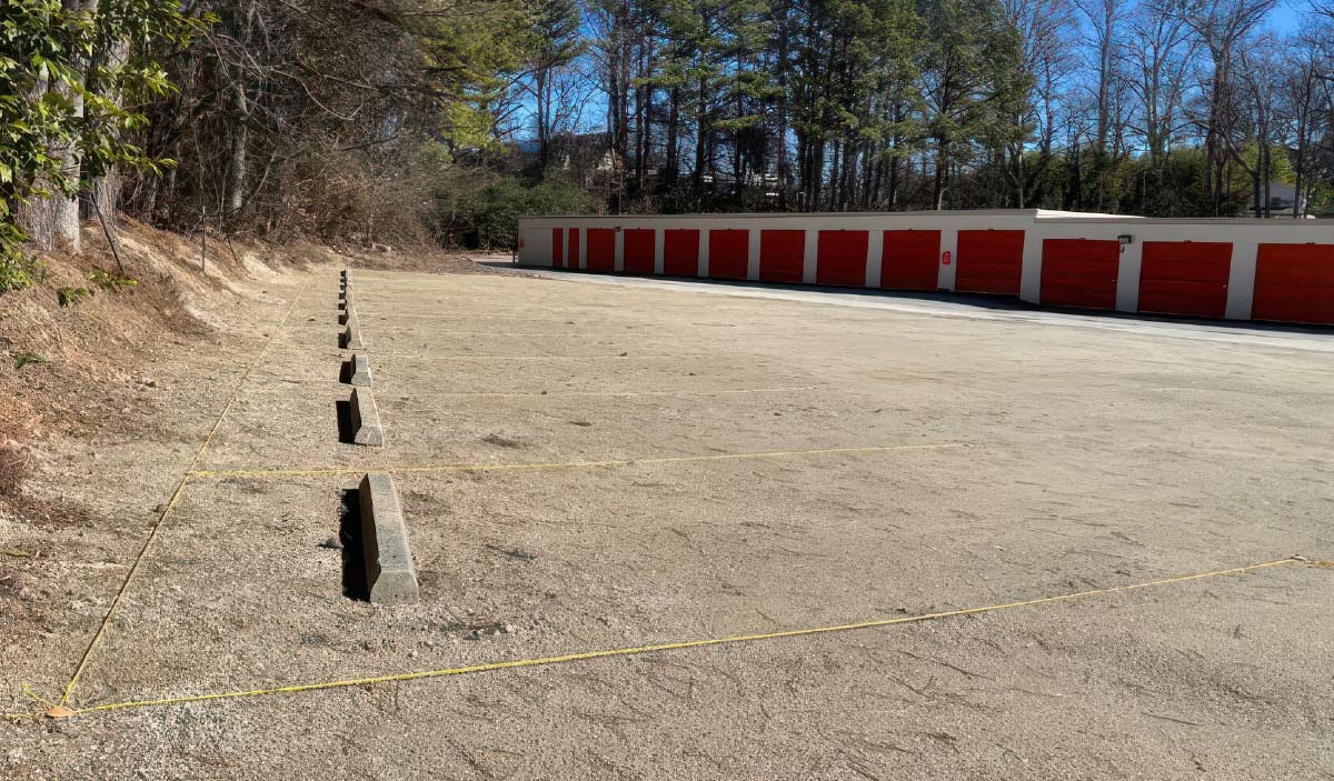 A storage facility parking area being prepared for paving, with concrete bumpers aligned along a yellow string marking out the spaces.