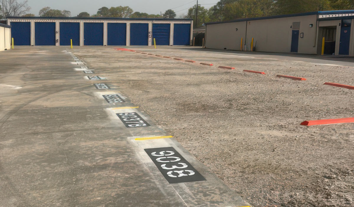 A completed asphalt parking lot at a storage facility featuring white painted space numbers and red parking bumpers aligned for use.