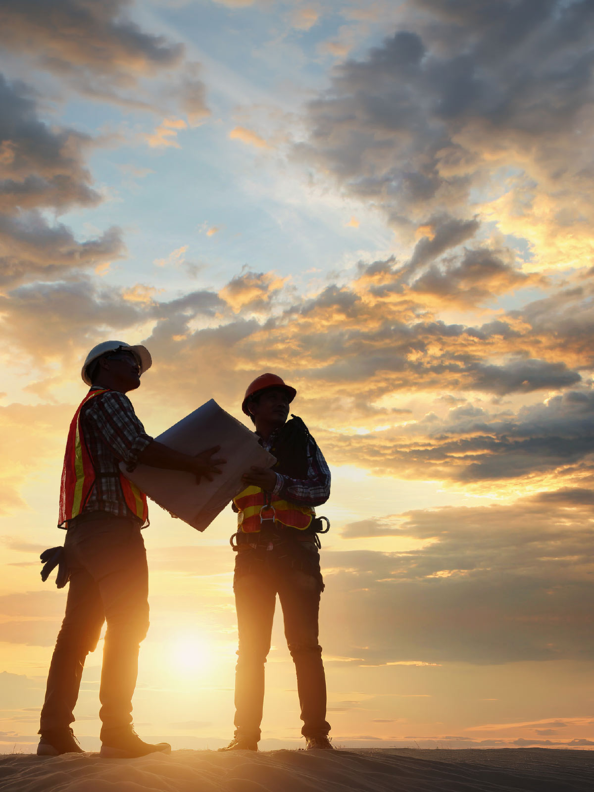 Silhouette of two construction workers in safety gear holding blueprints against a vibrant sunset sky.