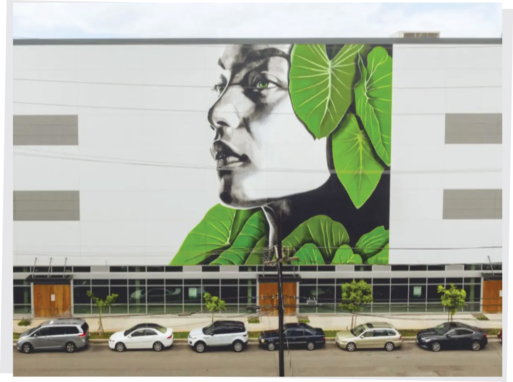 A large-scale exterior mural of a person's face framed by vibrant green leaves on a modern facility.