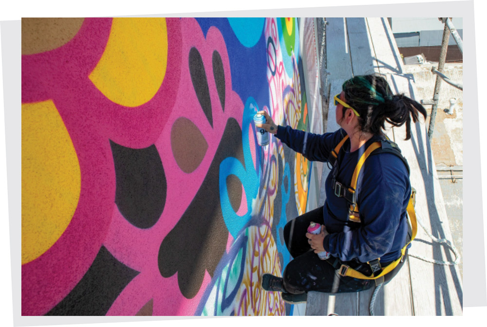 Close-up of an artist on a scaffold spray-painting a bright, multicolored geometric mural on a wall.