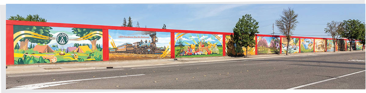 A long roadside retaining wall painted with a continuous series of colorful, historic community murals.