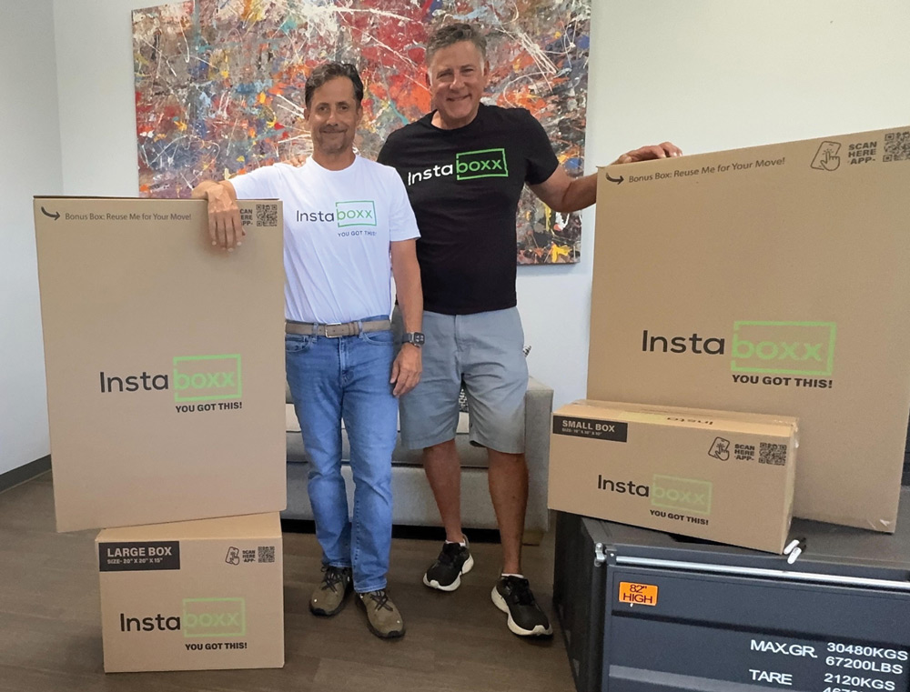 Two men standing next to cardboard boxes labeled "Instaboxx," with an abstract painting in the background.
