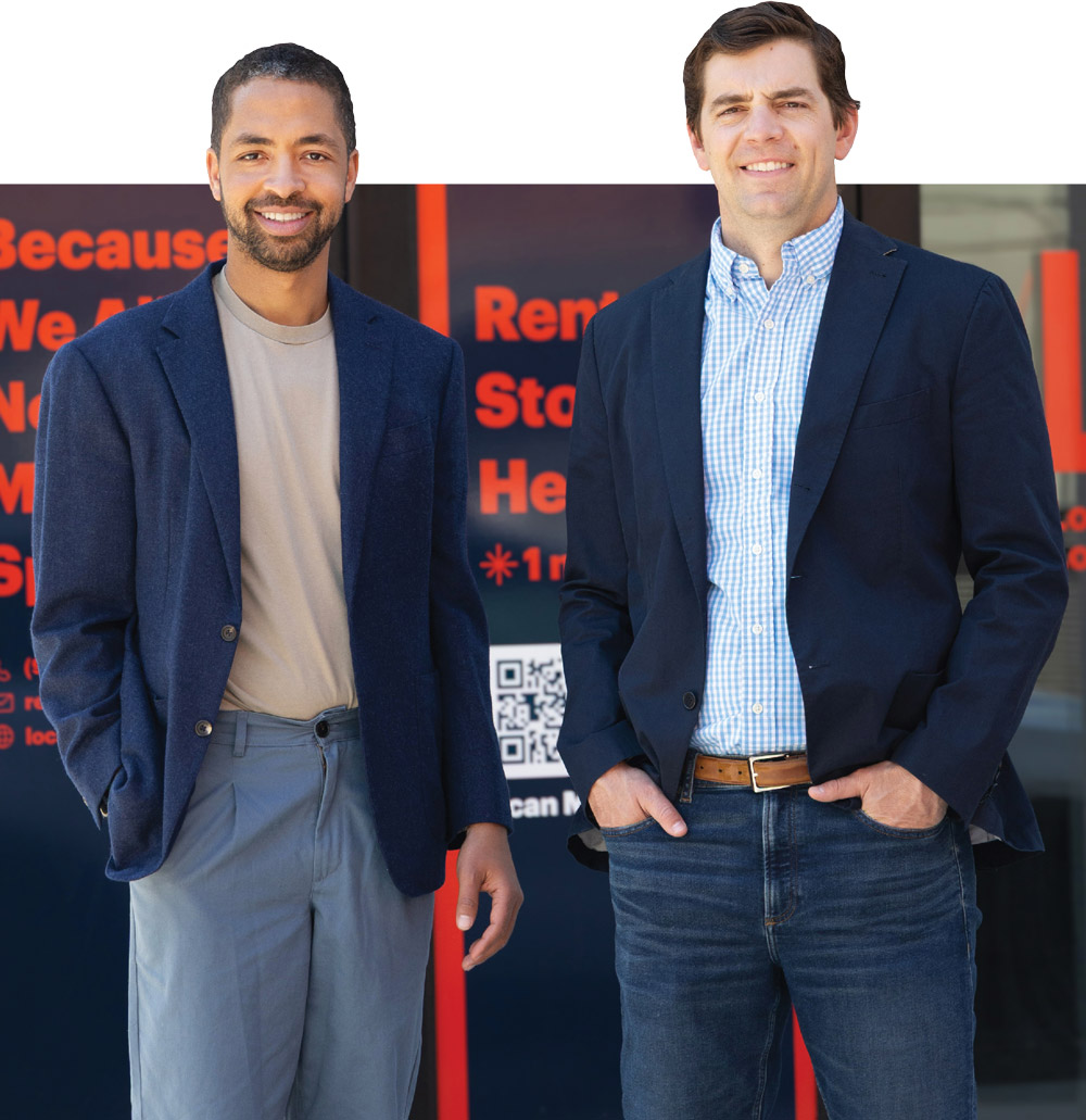 Two smiling businessmen wearing blazers stand next to each other in front of a sign with red lettering.