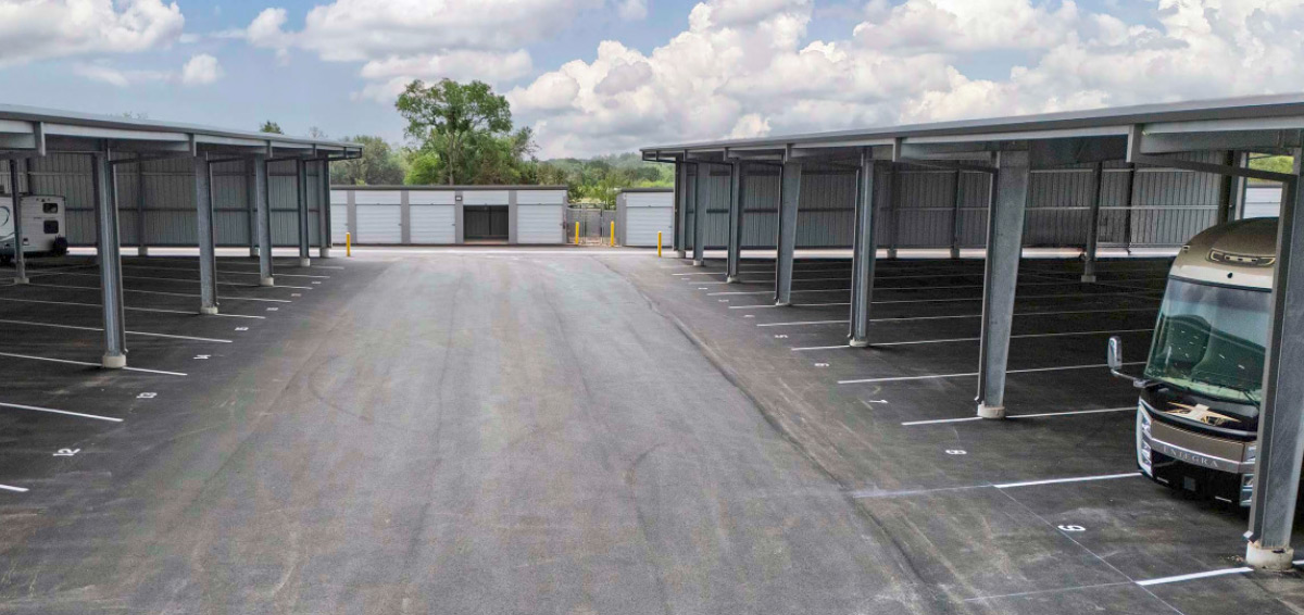 An outdoor driveway showing wide, covered parking bays designed for boats and RVs, with an Entegra motorhome parked in one stall.