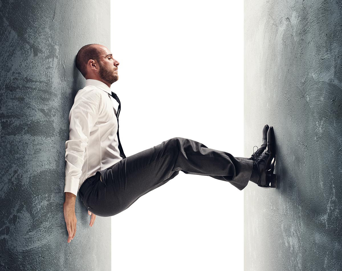 A man in a white shirt and tie suspends himself in mid-air between two textured grey walls. He braces his back against one wall and presses his feet against the other over a bright white background.