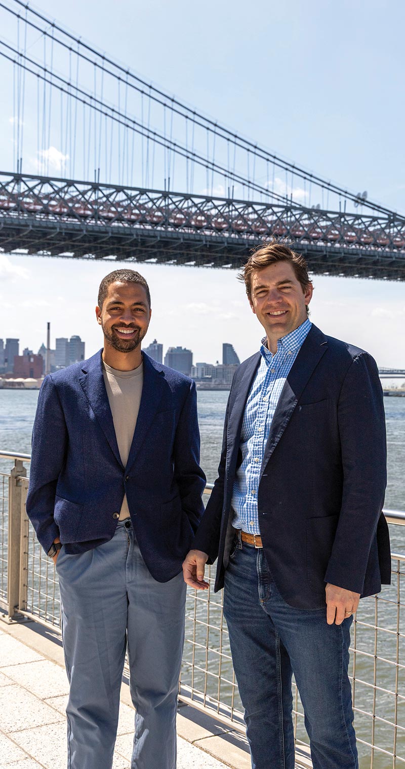 Alex Hartman and Peter Smyth standing outside on a waterfront walkway with a large suspension bridge and city skyline in the background.