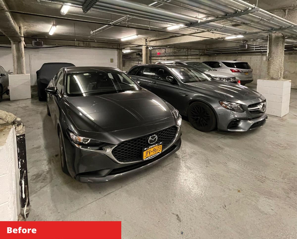Before conversion: Underground parking garage with concrete pillars and low ceilings, filled with several parked cars under fluorescent lighting.