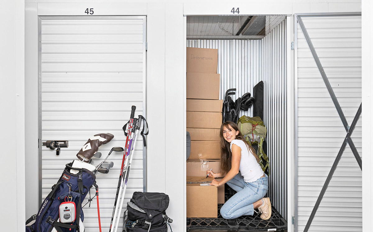 A customer kneels inside an open Local Locker storage unit filled with moving boxes and outdoor gear, next to another unit with golf clubs and skis outside.