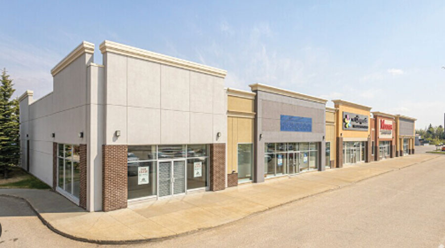Exterior view of a strip mall with several retail storefronts along a sidewalk.