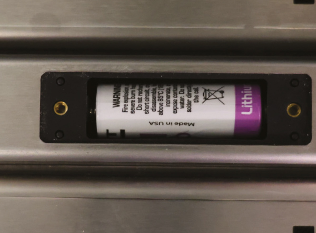 An open battery compartment on a metal smart latch housing a purple and white lithium battery made in the USA.