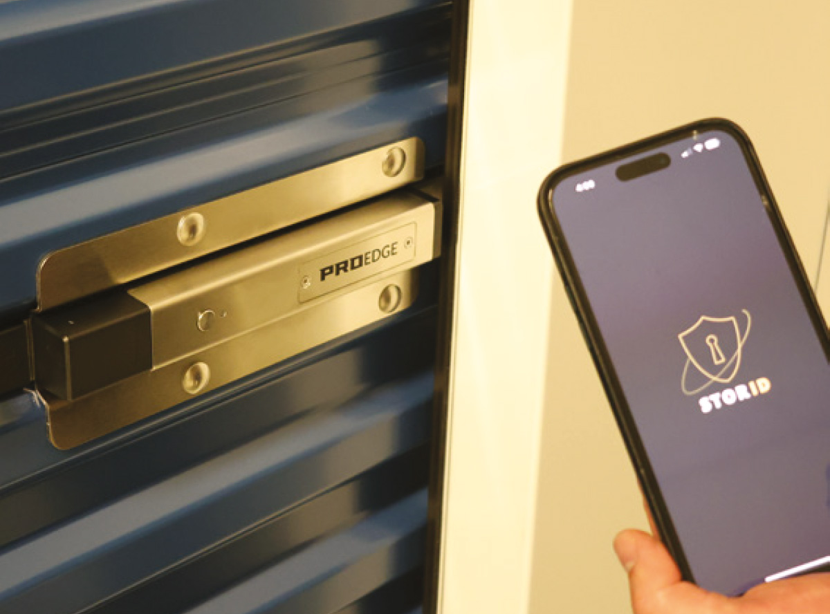 A hand holds a smartphone displaying the StorID app next to a ProEdge smart electronic security latch installed on a blue rolling door.