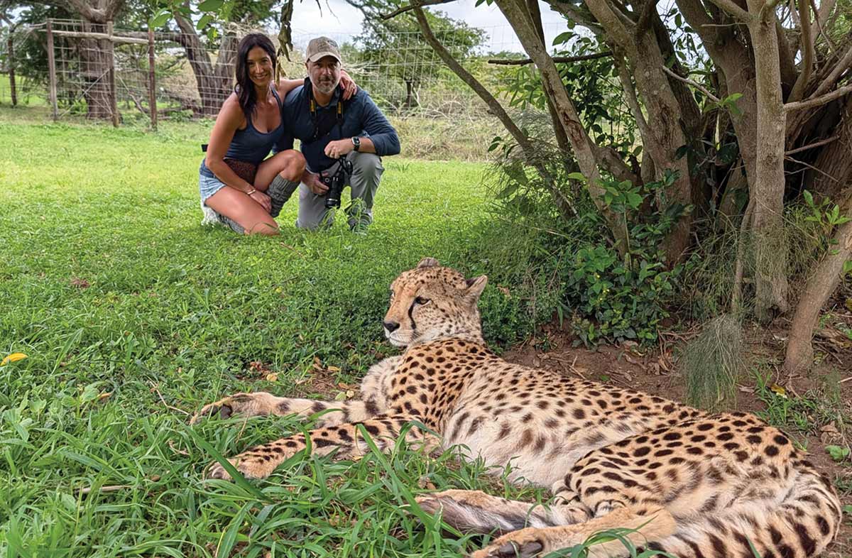 Renee and Matt Tulve observe a cheetah resting on an African safari as they are smiling crouched down in an open, grassy enclosure setting
