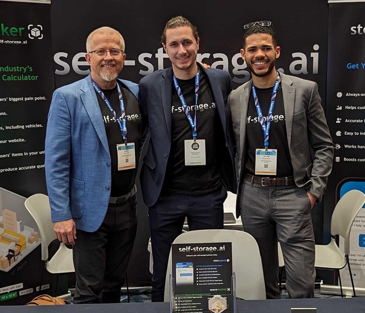 Three smiling colleagues pose in front of a self-storage.ai banner at a conference, wearing matching branded black shirts.