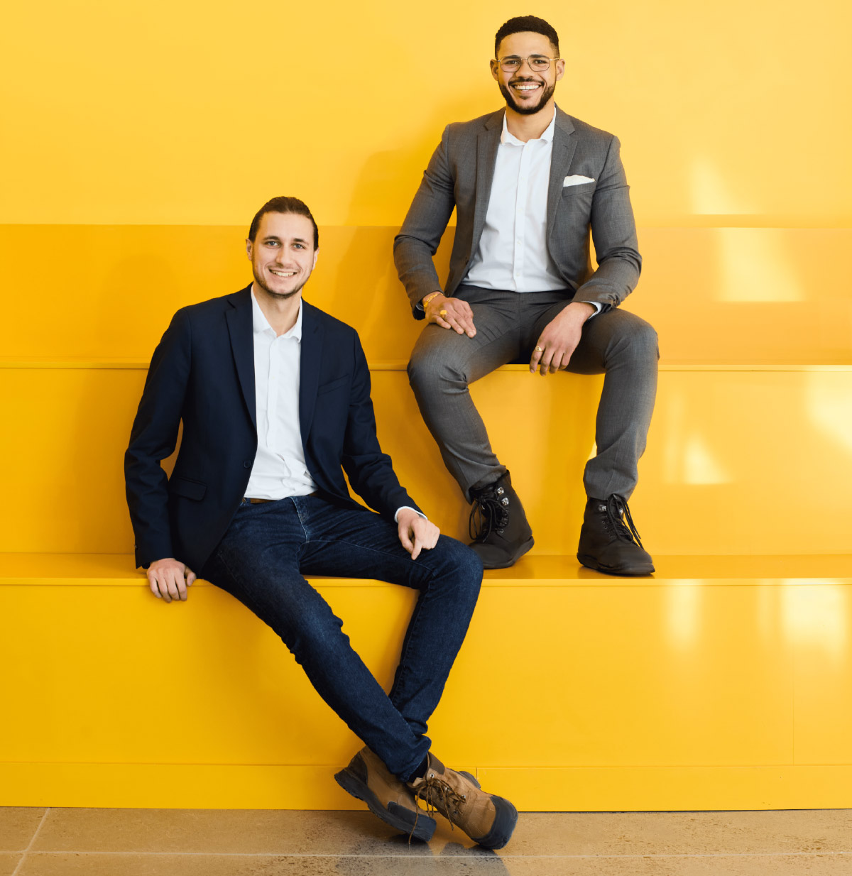 Two professionals in blazers and jeans sit on a bright yellow tiered bench, smiling for a corporate photo.