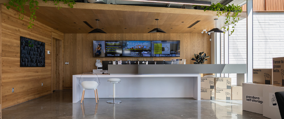 A sleek office lobby with wood panel walls, a long white desk, hanging lights, dynamic digital screens, and stacked moving boxes.