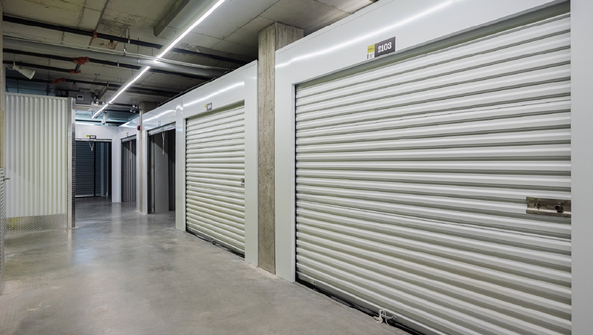 A brightly lit hallway inside a storage facility showing clean concrete floors and secure white roll-up storage unit doors.