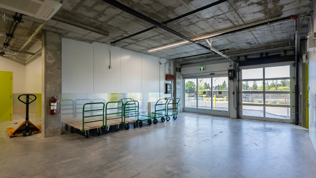 An indoor loading bay area featuring concrete floors, a wide garage door opening to the outside, a fire extinguisher, and green moving carts.