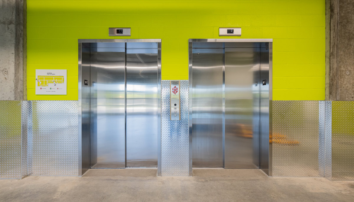 Two silver elevators set into a bright lime green accent wall, featuring diamond-plate metal wainscoting and a small floor map display.