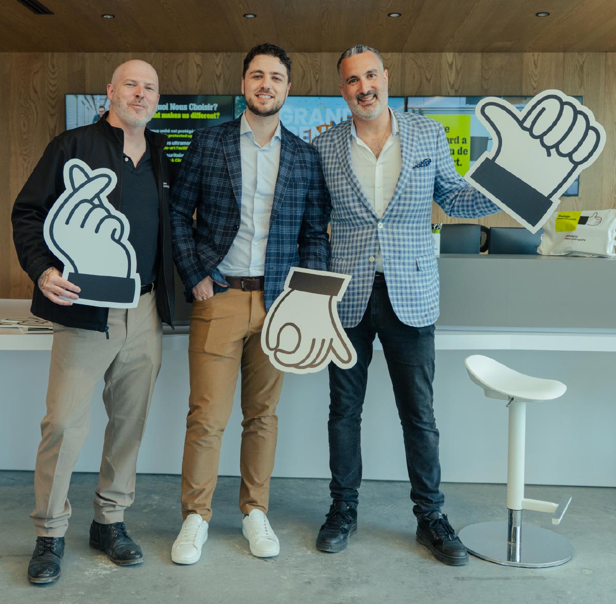 Three smiling men standing in a modern lobby holding playful, hand-shaped photo booth props featuring thumbs-up and finger-heart signs.