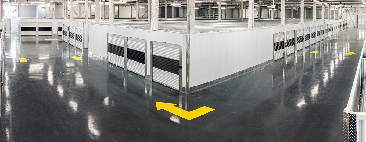 Large indoor drive-thru area with a polished dark floor marked with yellow traffic arrows. Rows of white storage units with roll-up doors extend down corridors beneath support pillars.