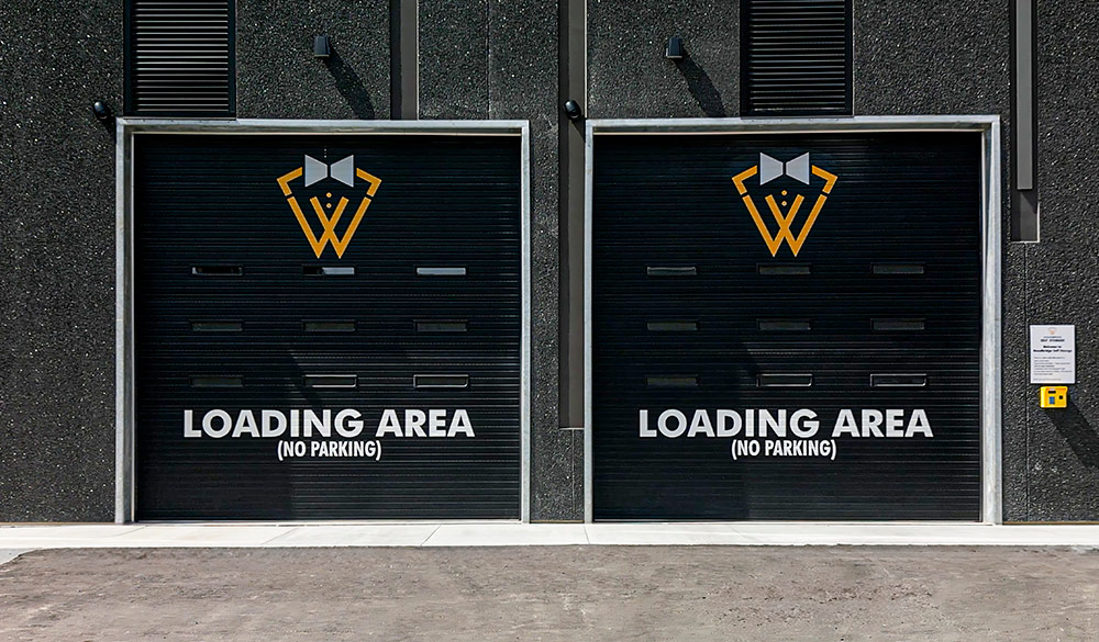 Two black roll-up garage doors on a dark textured wall. Each door features a yellow and white stylized "W" logo with a bowtie and white text reading "LOADING AREA (NO PARKING)."