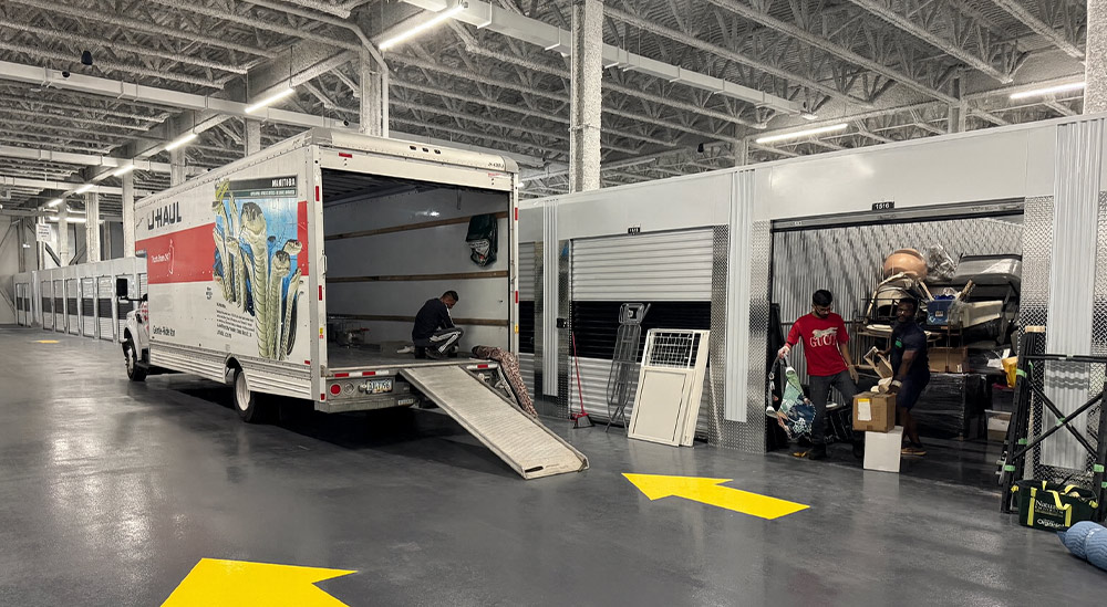 Indoor storage area where a U-Haul truck is parked with its ramp extended. One person works inside the truck while two others move boxes and household items into an open storage unit.