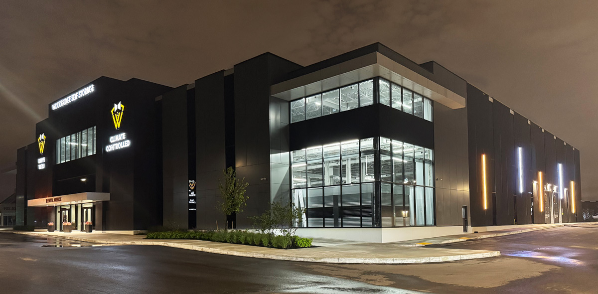 Exterior view of the modern Woodbridge Self Storage building lit up at night, showing bright interior lights through large glass windows, illuminated signage, and vertical light accents.
