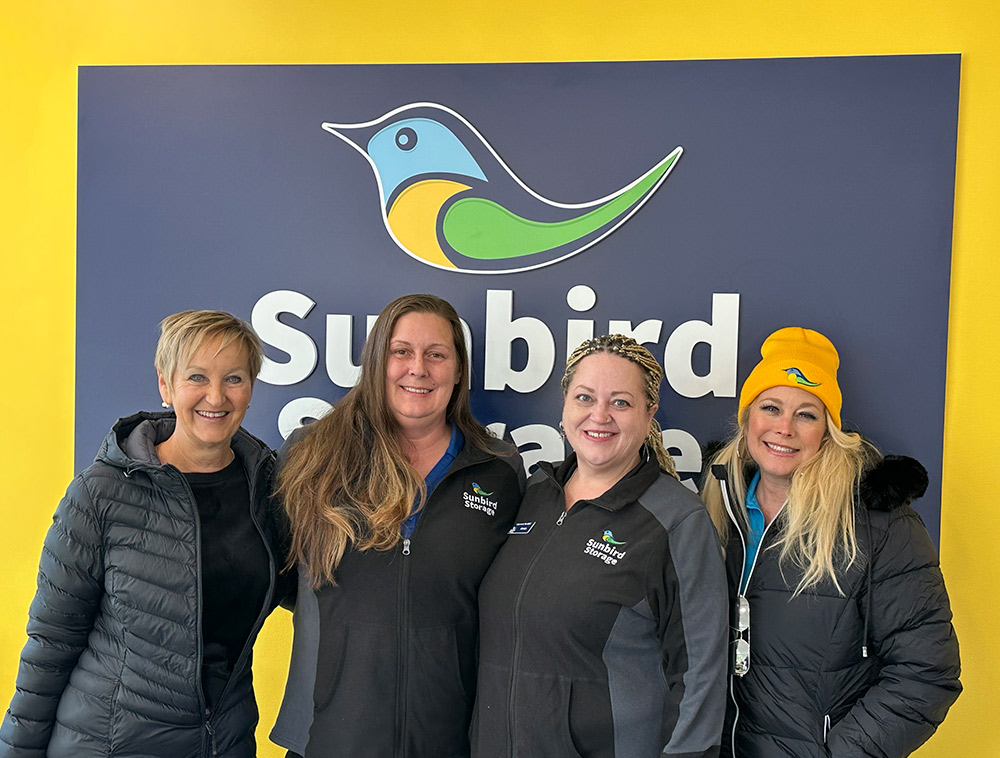 Four women from the Sunbird Storage team smile in front of a blue company sign with a bird logo on a yellow wall. Two women wear black company jackets, and one wears a yellow beanie.