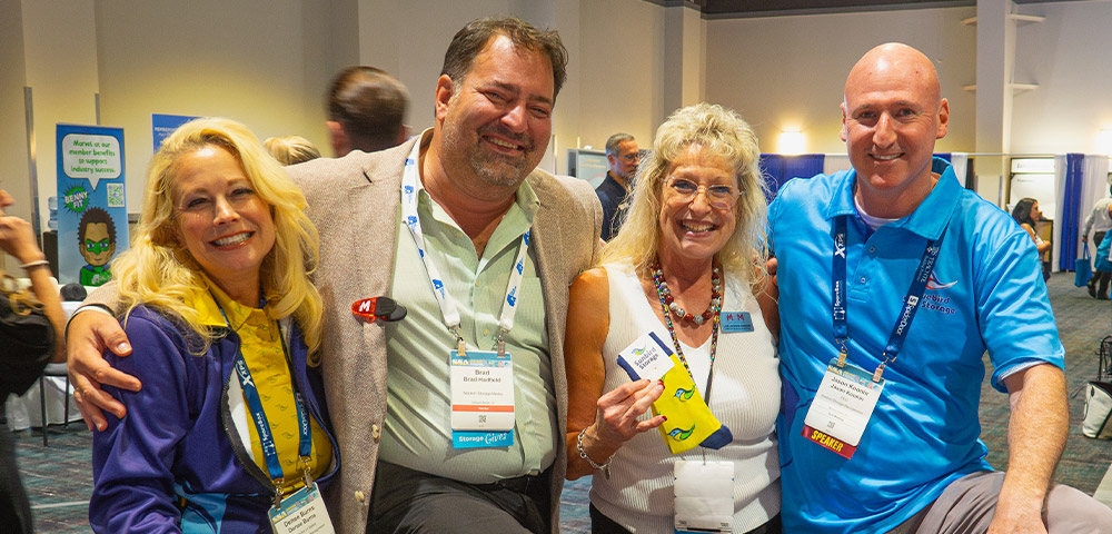 Denee Burns, Brad Hadfield, Lauri Longstrom-Henderson, and Jason Koonin smile at a conference. Lauri holds a pair of yellow socks with a bird logo, and three people wear event lanyards.