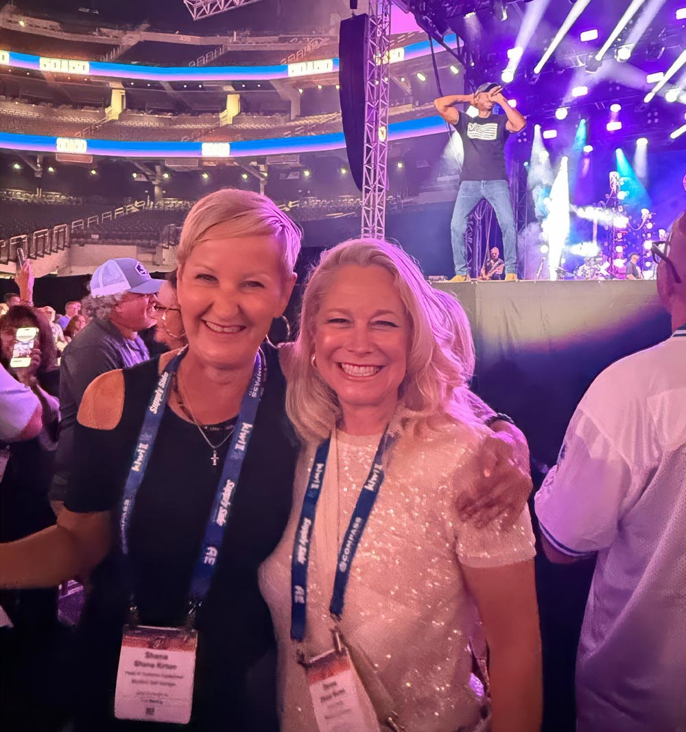 Shana Kirton and Denee Burns smile together while wearing conference lanyards. They are inside a large, dimly lit arena with a performer on a brightly lit stage in the background.
