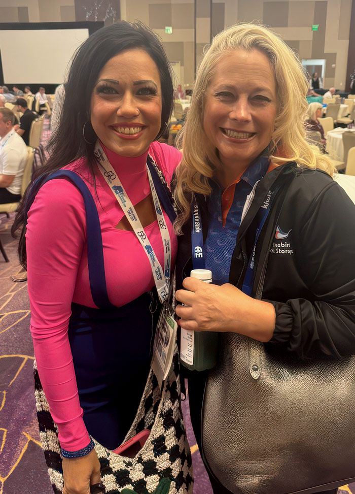 Jessica Johnson and Denee Burns pose together indoors wearing lanyards. Jessica wears a pink shirt, and Denee wears a black jacket with a bird logo and holds a beverage bottle.