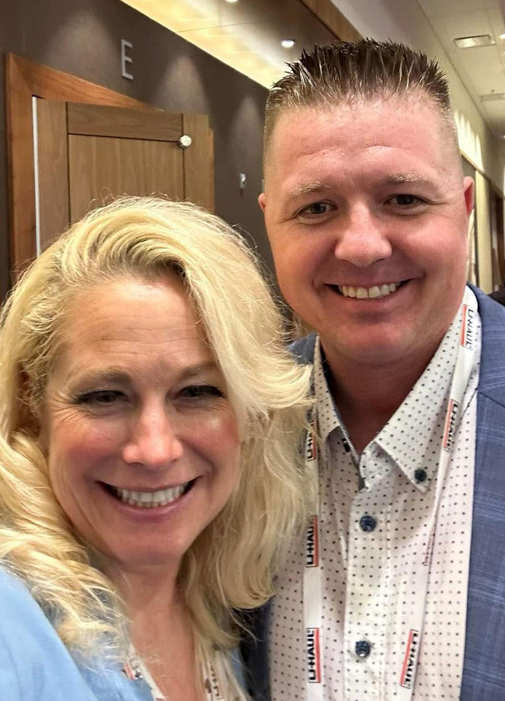 Close-up photo of Denee Burns and Dusty Lucy smiling indoors. Dusty wears a patterned white shirt, a blue blazer, and a U-Haul lanyard.