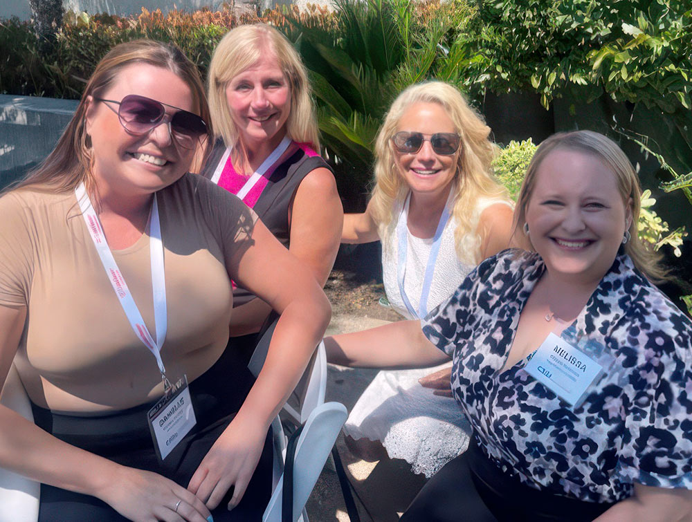 Danielle Pancheri, Christina Powell, Denee Burns, and Michelle Wight-Sands smile while seated outdoors in white chairs on a sunny day with green foliage behind them.