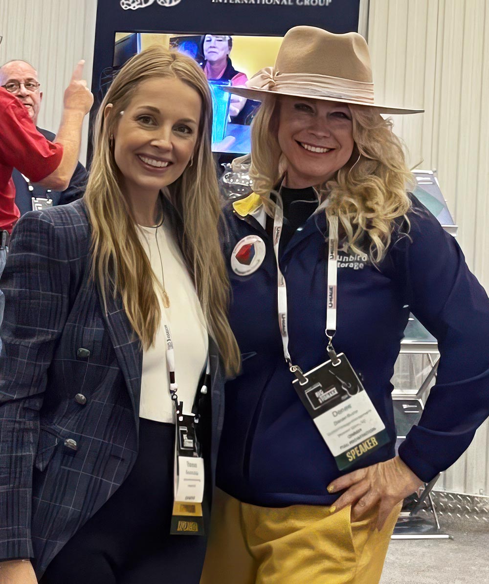 Theresa Gallas and Denee Burns smile together at an event. Theresa wears a plaid blazer, and Denee wears a tan wide-brimmed hat and a blue jacket with a speaker badge.
