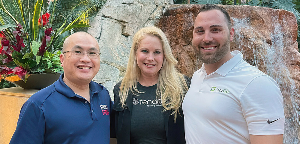 Denee Burns stands between two men, including James Reid on the right, in front of an indoor rock waterfall feature. All three are smiling and wearing corporate logo polo and t-shirts.
