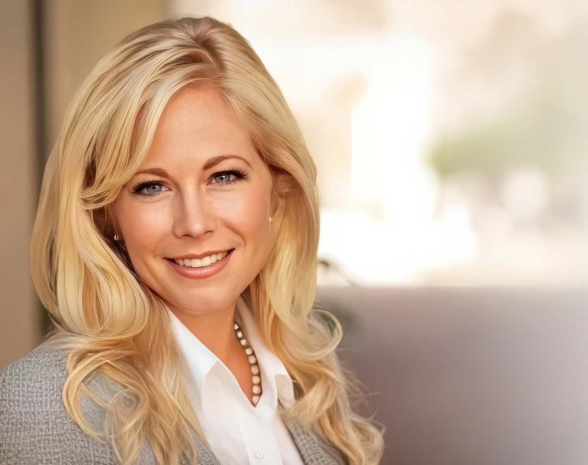 Headshot of Denee Burns smiling directly at the camera. She has long blonde wavy hair and blue eyes, and she is wearing a white collared shirt, a pearl necklace, and a gray blazer against a softly blurred background.