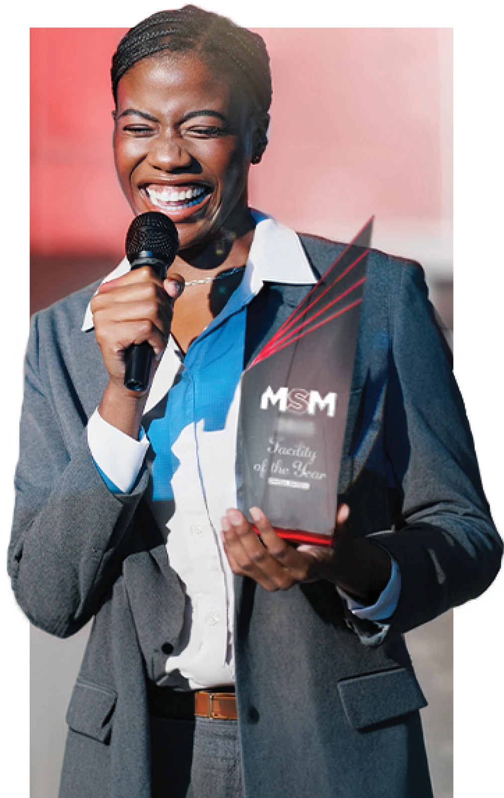 Person in a suit holding a microphone and a trophy, smiling outdoors.
