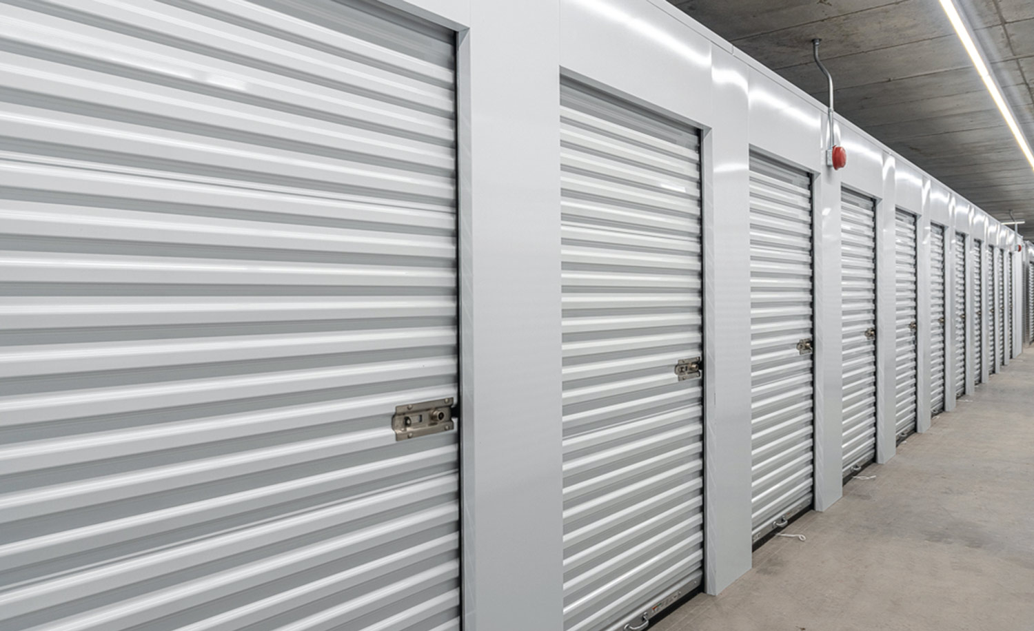 A long, clean indoor corridor lined with bright white corrugated roll-up doors for self-storage units.