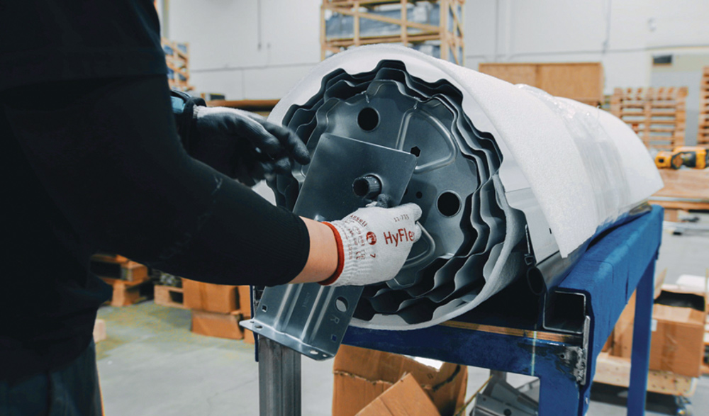 A factory worker wearing safety gloves installs hardware onto the end of a rolled-up metal storage door sheet.