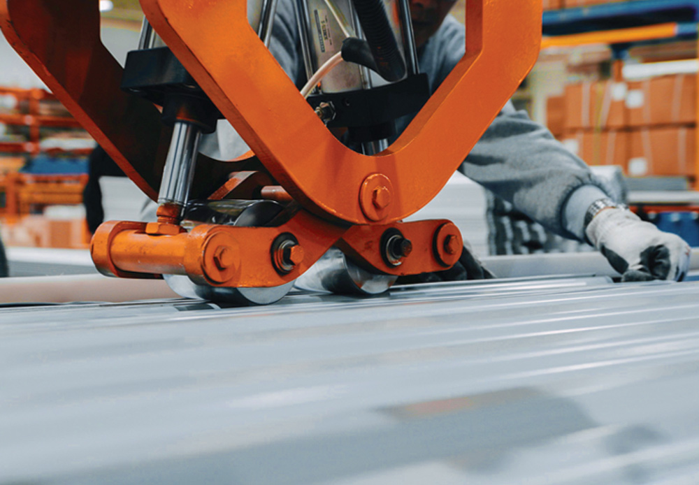 Heavy-duty orange machinery presses down on a sheet of corrugated silver metal during manufacturing.