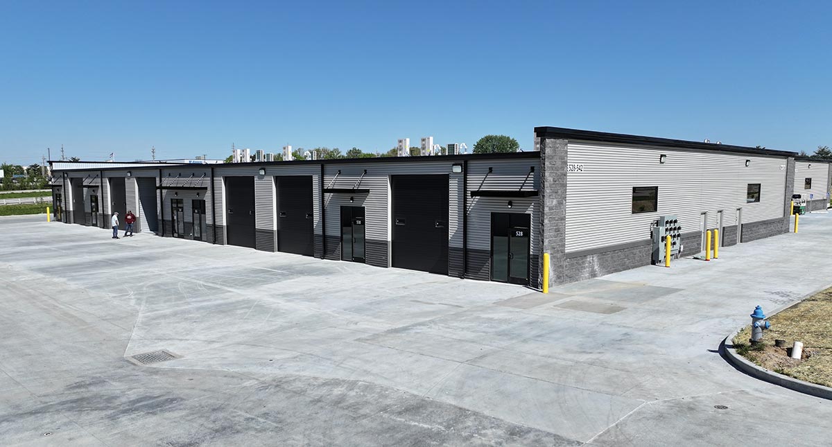A modern commercial warehouse building with large black roll-up doors and concrete parking.