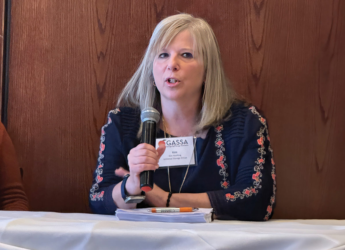 Kim Hoelting speaks into a microphone as a Georgia Self Storage Association panelist while seated behind a table draped with a white cloth