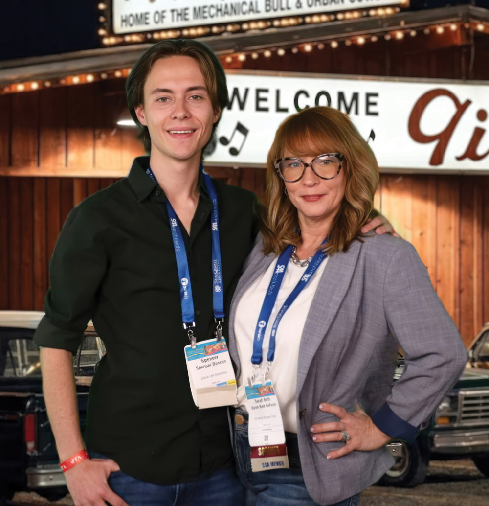 A smiling young man and a woman wearing conference lanyards pose together in front of a rustic, lit-up country western venue.