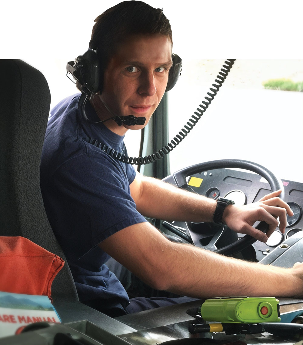 A man wearing a headset sits in the driver's seat of a large vehicle, holding the steering wheel and looking at the camera.